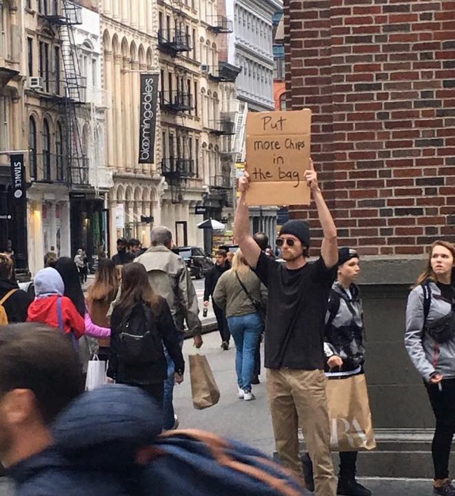The meme features a person holding a sign in a crowded city street, with a humorous message. The sign reads "Put more chips in the bag" in a playful, exasperated tone, implying that the person is frustrated with the lack of snacks in their bag. The image is likely intended to be humorous and relatable, poking fun at the common experience of running out of snacks.